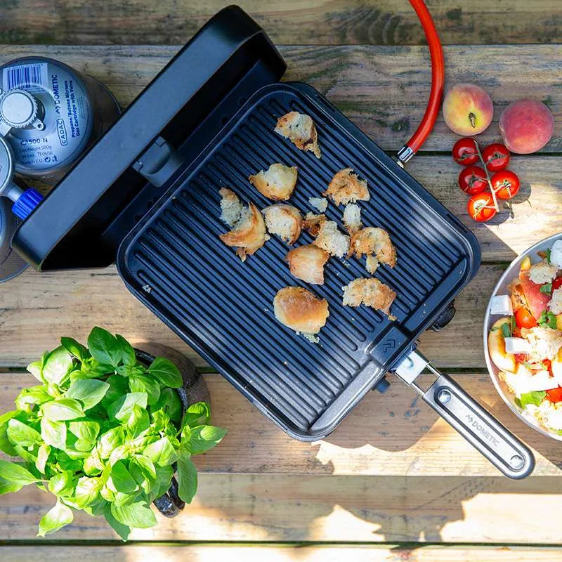 Black camping stove with food on a wooden table with vegetables and a salad.