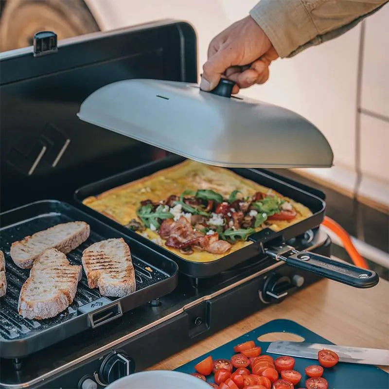 Person cooking on a portable grill with an omelette and bread, surrounded by kitchen items.