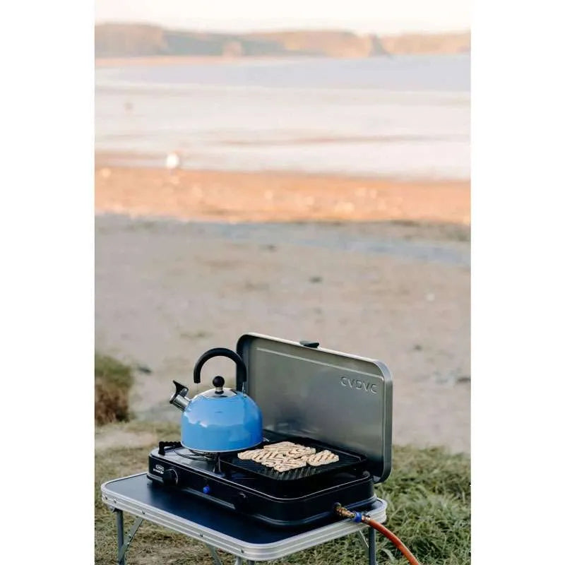 Portable grill with a blue kettle and food on a camping stove outdoors.