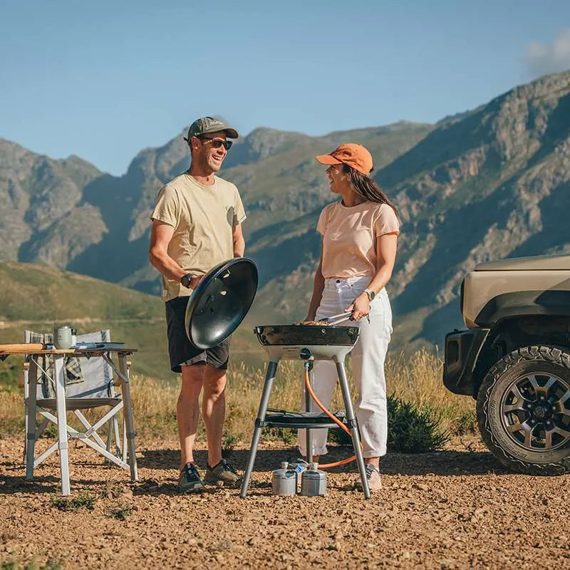 Two people preparing food on a portable grill in a mountainous outdoor setting.