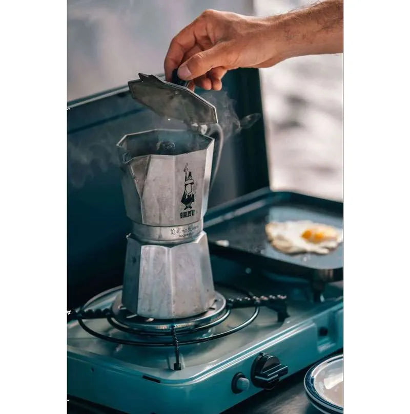Person using a Moka pot on a camping stove with a blurred background