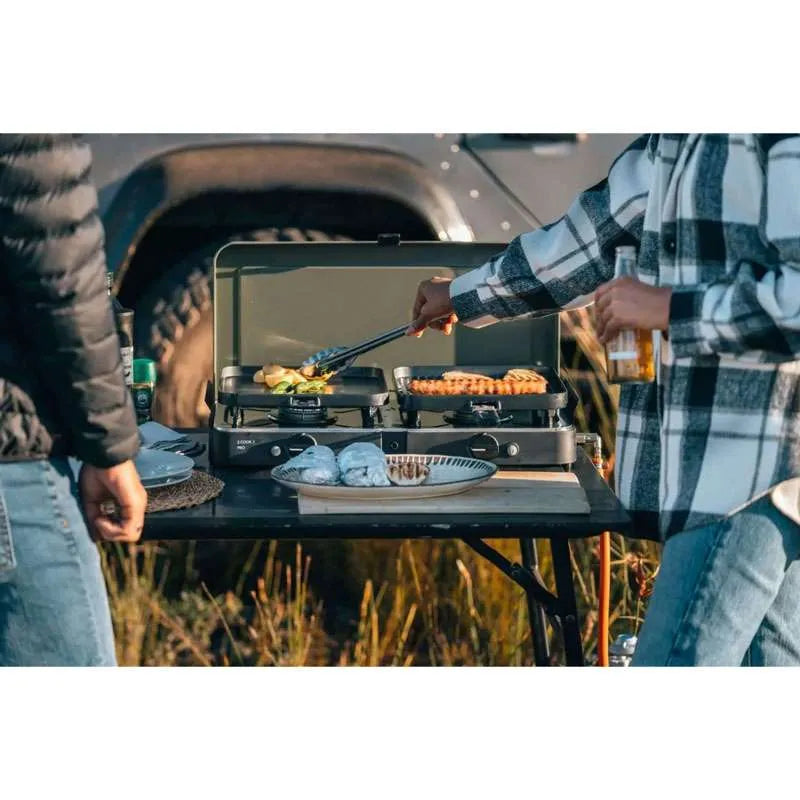 Two people grilling outdoors with a portable stove and food items.