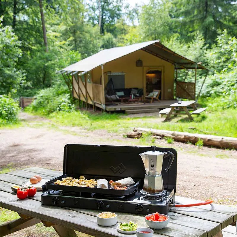 Camping setup with a portable stove, coffee maker, and food on a wooden table in front of a tent in a forest.