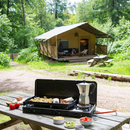 Camping setup with a portable stove, coffee maker, and food on a wooden table in front of a tent in a forest.