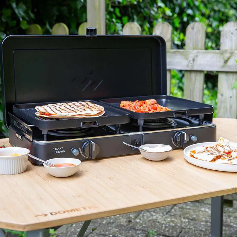 Portable gas grill on a wooden table with food being prepared outdoors.
