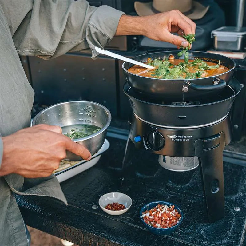 Person cooking outdoors using a portable stove with food ingredients nearby.