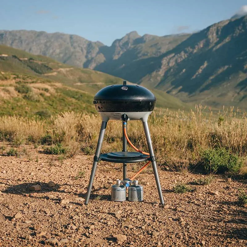 Portable barbecue grill on a stand with two propane tanks in a mountainous landscape.