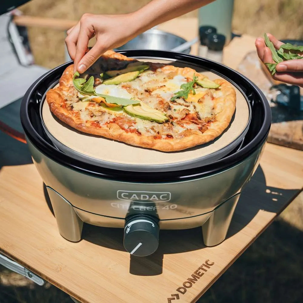 Person preparing a pizza on a portable Cadac grill outdoors.